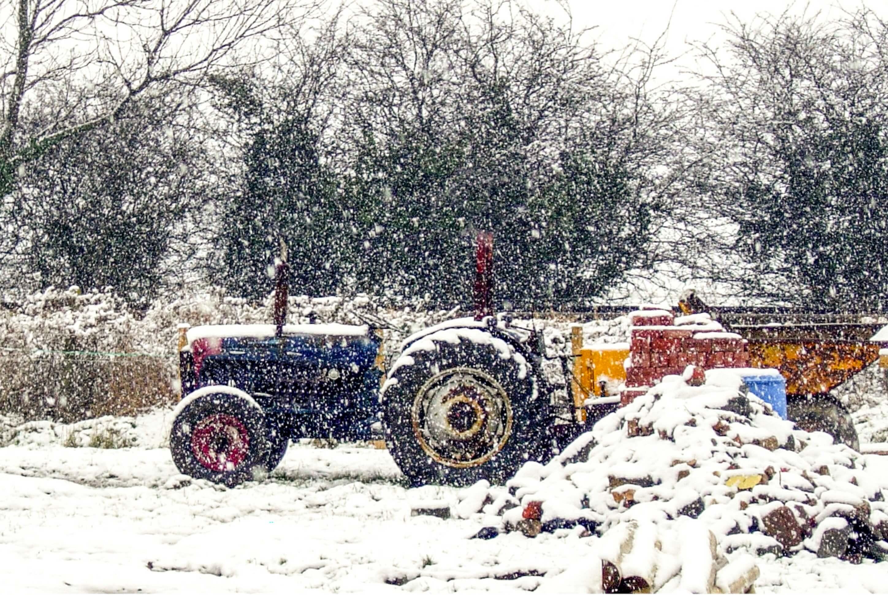 Heavy snow is falling. A blue and red tractor with bright yellow trailer is parked, a pile of logs sits in the foreground