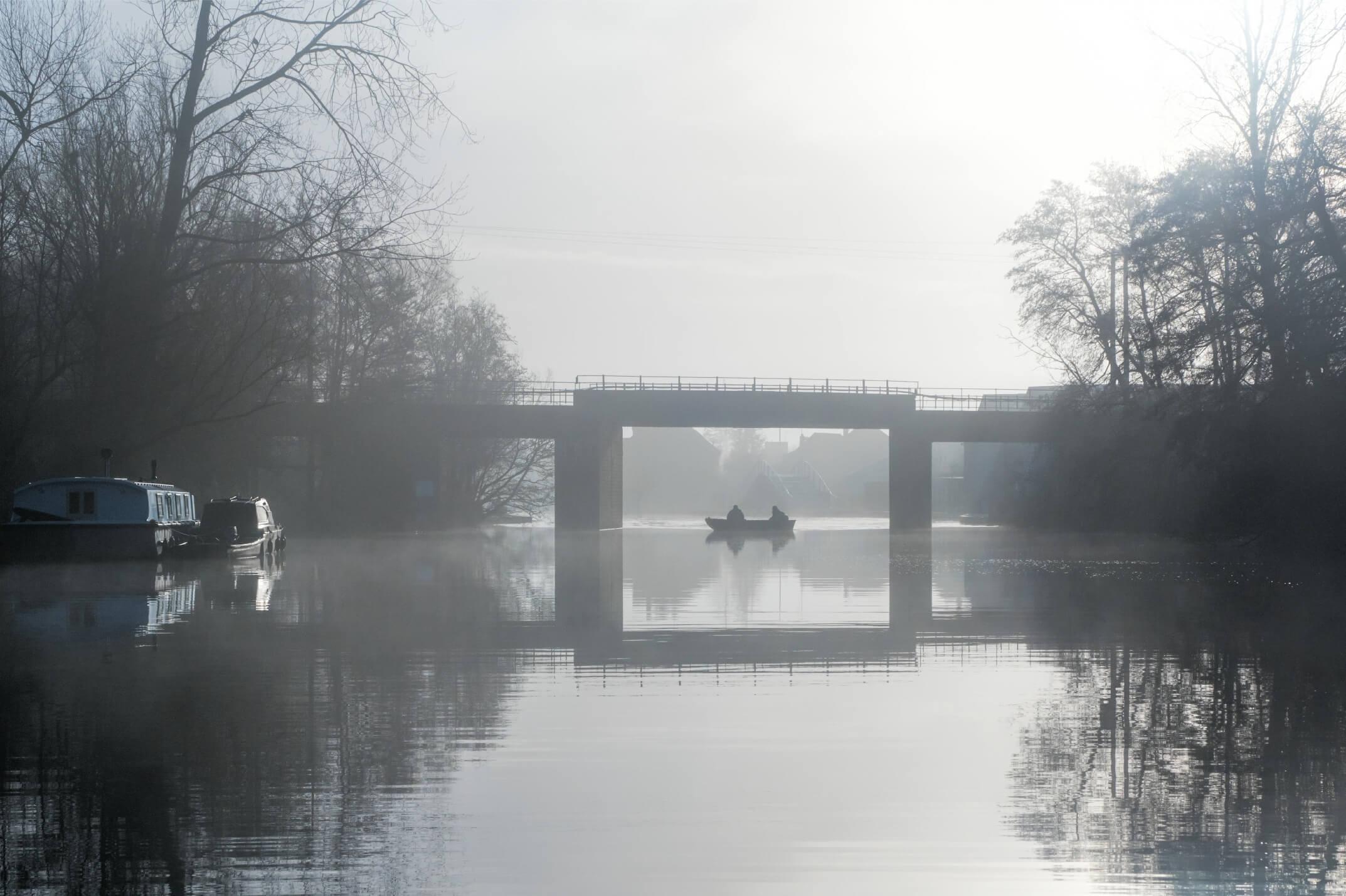 A misty morning on the river. A small fishing boat comes into view under Wroxham railway bridge. As the sun breaks through a reflection of the bridge frames the fishing boat.