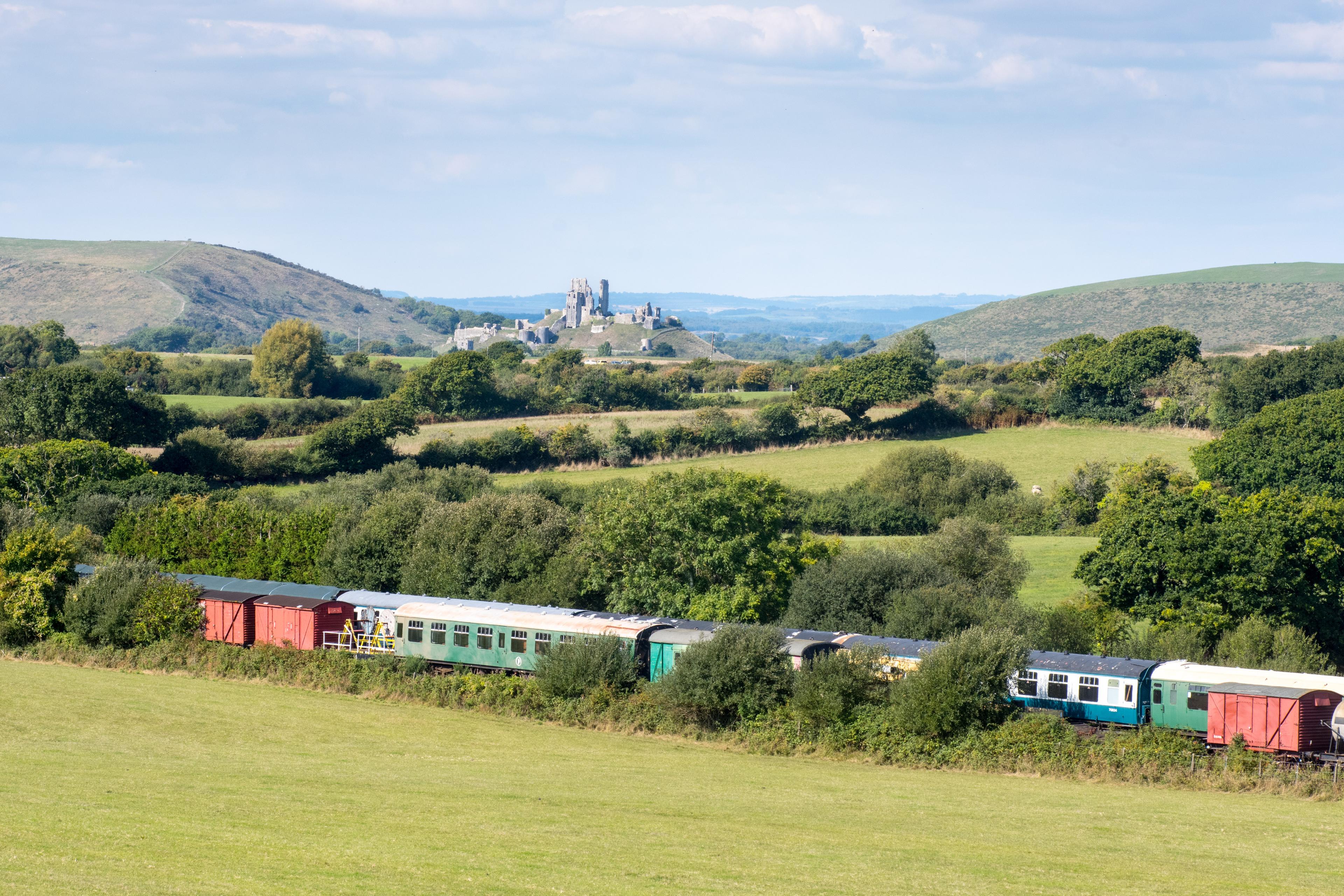 Hills and green fields, in the foreground a steam train rests and in the distance is the ruins of a castle on a hill.