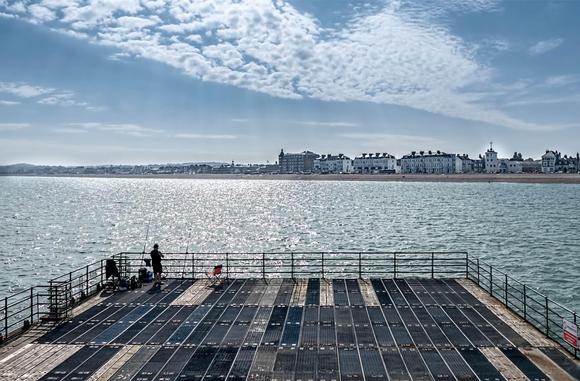 Looking across the sea from the end of a pier, a lone person is fishing.