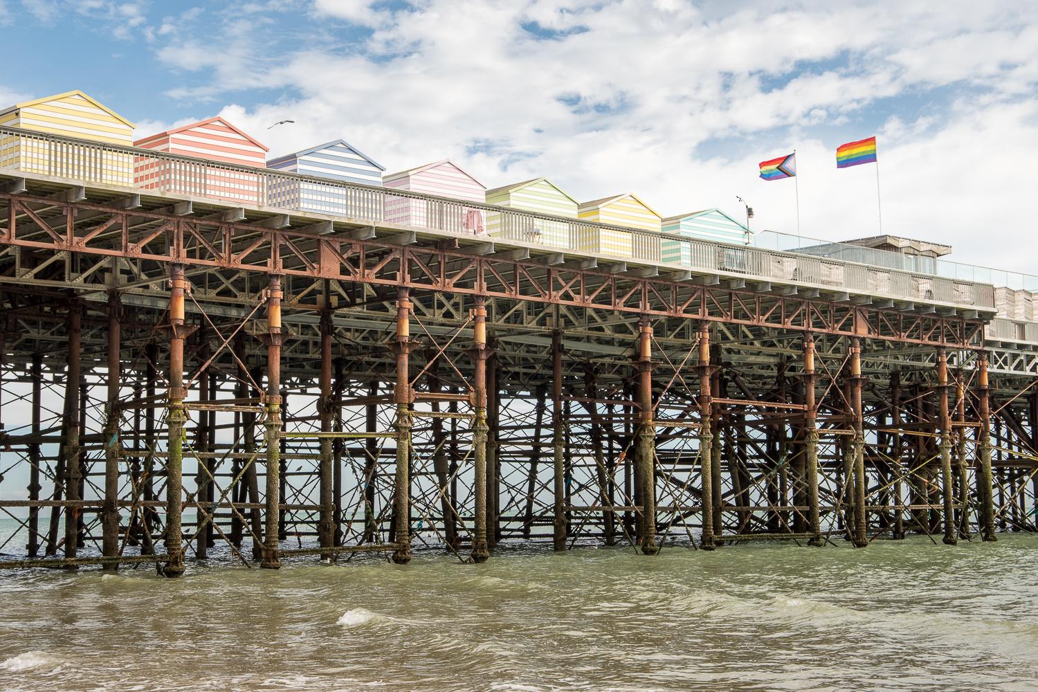 Multi-coloured beach huts sit on a pier. Underneath the pier issupporting ironwork entangled with seaweed and fishing paraphernalia