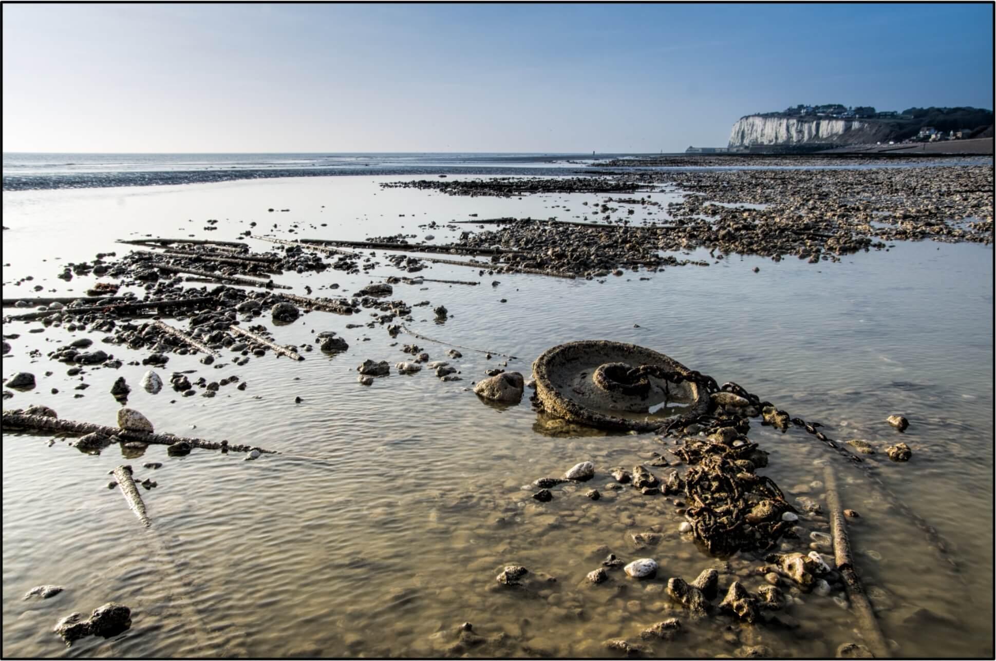 Exposed rusted train wheel attached to chains, encrusted with limpets and mussels, across the bay is a chalk cliff.