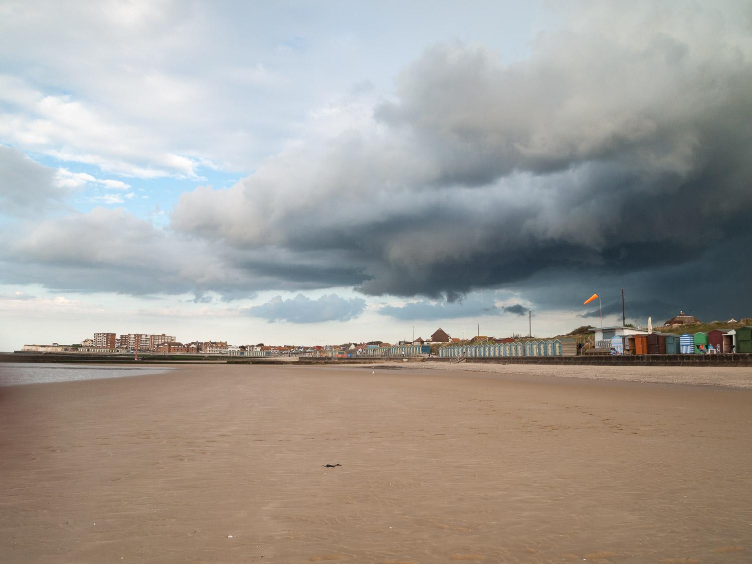 Single ominous black rain cloud sweeping across beach huts and sandy bay, sweeping out to sea.