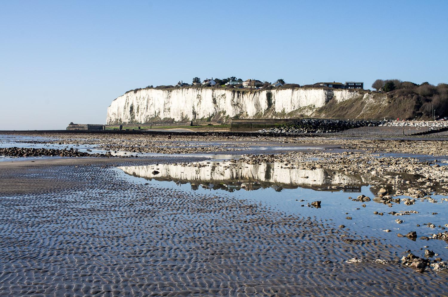 Low tide, blue skies, a reflection of Kingsdown cliffs in a rock pool, a distant figure sits alone on the chalk rocks