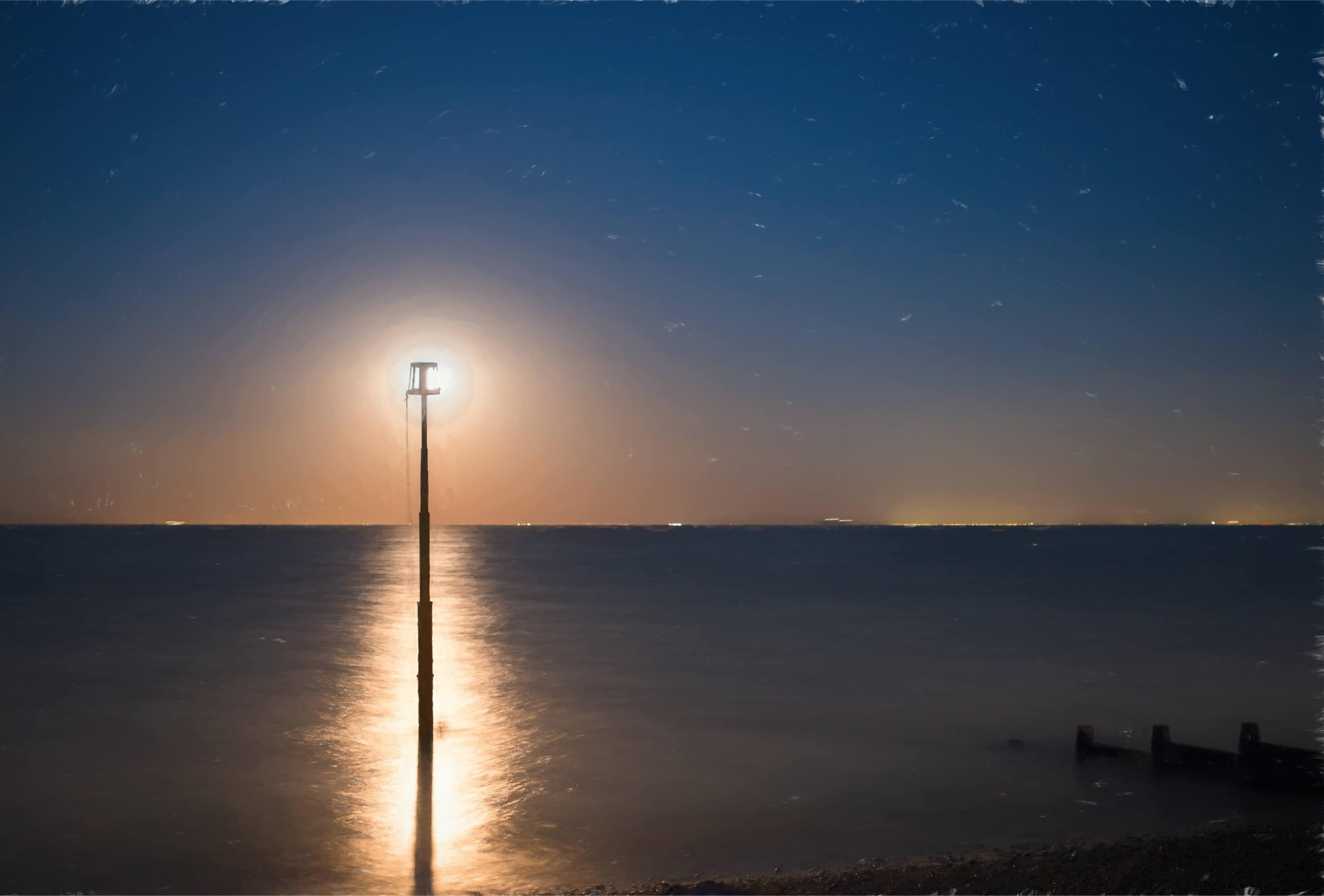 The full moon creates a path of moonlight across a calm sea, the distant orange glow of Calais is on the horizon